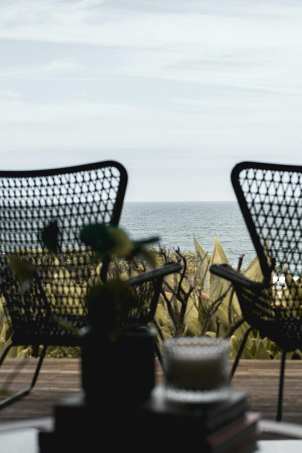 Wooden terrace with wicker armchairs placed nearby lush greenery in front of endless rippling seawater